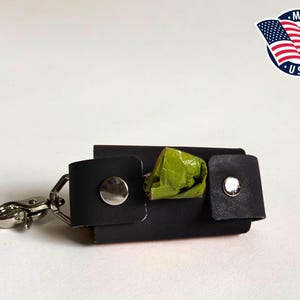 May include: A black rectangular waste bag dispenser featuring two silver-toned metal snap buttons and a silver-toned metal swivel lobster clasp. A small portion of a green plastic waste bag is visible protruding from the center of the dispenser.