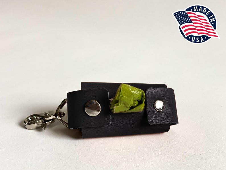 May include: A black rectangular waste bag dispenser featuring two silver-toned metal snap buttons and a silver-toned metal swivel lobster clasp. A small portion of a green plastic waste bag is visible protruding from the center of the dispenser.