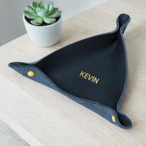 以下が含まれることがあります： ゴールドの文字で「KEVIN」と書かれた、濃い青色の革製小物入れトレイ。トレイは角が丸い三角形の形で、黄色のスナップ留め具で固定されています。小さな鉢植えの多肉植物が近くにあります。