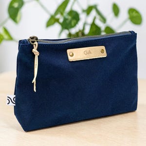 May include: A navy blue canvas pouch with a gold zipper and a leather tag that reads "GA". The pouch has a small white tag on the side and a gold tassel attached to the zipper. The pouch is sitting on a light wood surface.