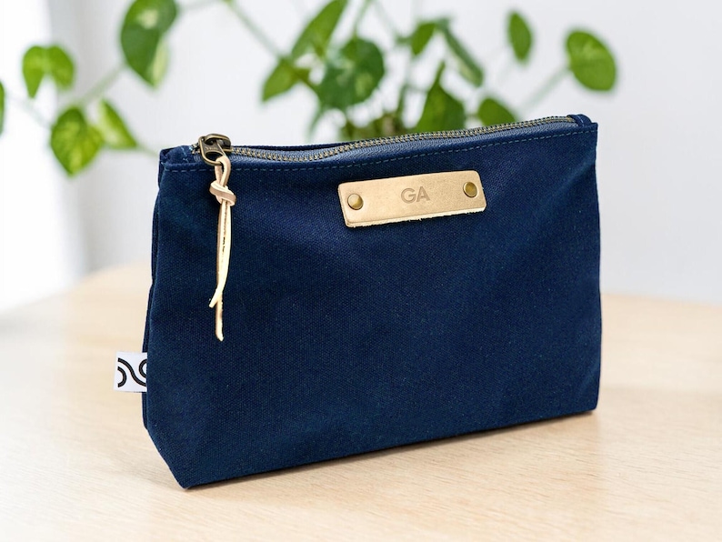 May include: A navy blue canvas pouch with a gold zipper and a leather tag that reads "GA". The pouch has a small white tag on the side and a gold tassel attached to the zipper. The pouch is sitting on a light wood surface.
