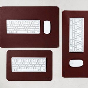 May include: A collection of three burgundy-colored desk mats with white keyboards and mice. The mats are rectangular, with rounded corners. The keyboards and mice are positioned on the mats, ready for use. The setup is clean and modern.