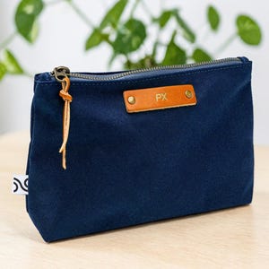 May include: A navy blue canvas pouch with a brass zipper and a leather pull. The pouch features a small, rectangular leather tag with the initials "PX" embossed on it. A small white tag with a black design is sewn on the side.