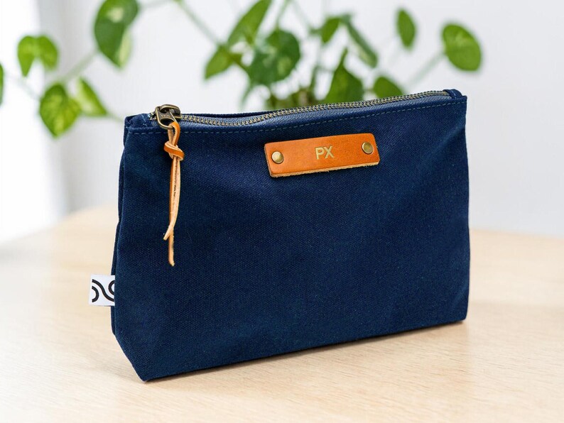 May include: A navy blue canvas pouch with a brass zipper and a leather pull. The pouch features a small, rectangular leather tag with the initials "PX" embossed on it. A small white tag with a black design is sewn on the side.