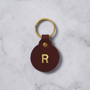 Puede incluir: Un llavero redondo de cuero marrón oscuro con un llavero dorado. El llavero presenta la letra dorada "R" y un pequeño remache dorado. El fondo es una superficie de mármol blanco y gris.