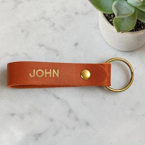 May include: A tan leather keychain featuring the name JOHN embossed in capital letters. The strap is secured with a gold-toned metal rivet and includes a matching circular gold-toned metal split ring for attaching keys or other small items.
