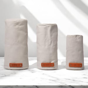 May include: Three cylindrical beige fabric covers of varying heights, each featuring a rectangular tan leather tab at the base secured with two brass rivets. Each leather tab is embossed with the letters LK in a simple, clean font.