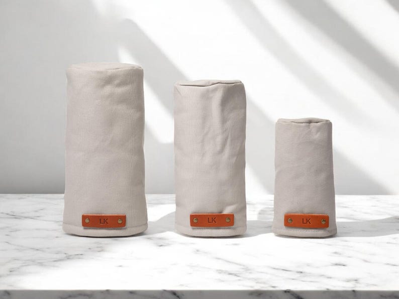 May include: Three cylindrical beige fabric covers of varying heights, each featuring a rectangular tan leather tab at the base secured with two brass rivets. Each leather tab is embossed with the letters LK in a simple, clean font.