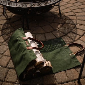 May include: A green canvas log carrier with brown leather handles holds several logs. The carrier is open, displaying the wood. A metal fire pit with a burning fire is in the background.