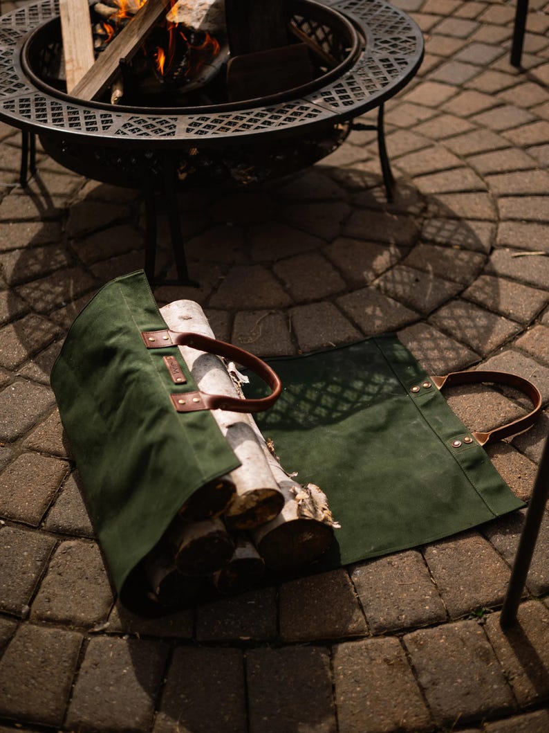 May include: A green canvas log carrier with brown leather handles holds several logs. The carrier is open, displaying the wood. A metal fire pit with a burning fire is in the background.