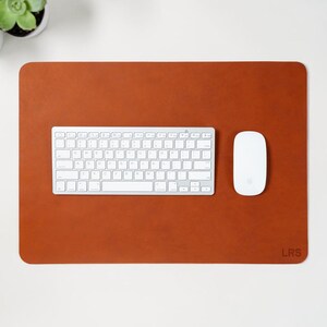 May include: A rectangular, rust-colored leather desk mat with rounded corners. A white keyboard and mouse are centered on the mat. The initials "LRS" are in the lower right corner. Two small potted succulents are in the upper left corner.