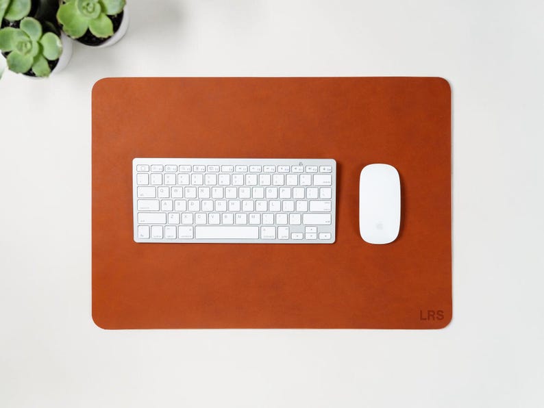 May include: A rectangular, rust-colored leather desk mat with rounded corners. A white keyboard and mouse are centered on the mat. The initials "LRS" are in the lower right corner. Two small potted succulents are in the upper left corner.