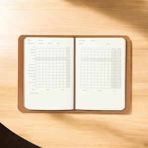 May include: Open notebook with grid pages, inside a brown leather cover, resting on a light wood surface. The notebook is open, revealing two facing pages with a grid pattern for tracking or journaling. The leather cover has a simple, clean design.