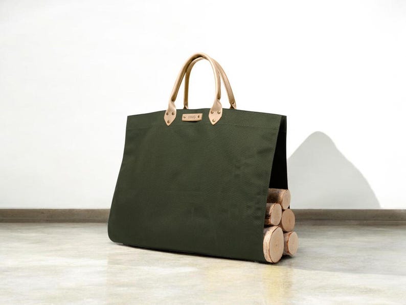 May include: An olive green canvas tote bag with tan leather handles and accents. The bag is filled with several cut logs. The bag is designed for carrying firewood.