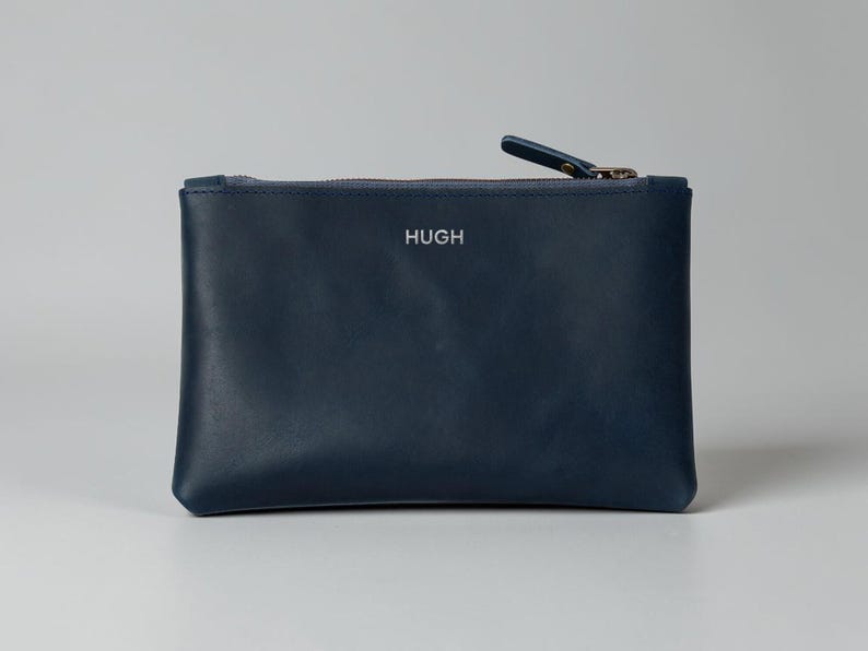 Pode incluir: Uma bolsa de couro azul marinho com fecho de correr e uma pequena aba. A palavra "HUGH" est&aacute; gravada em prata na frente. A bolsa &eacute; retangular e parece estar vazia. Ideal para guardar pequenos objetos.