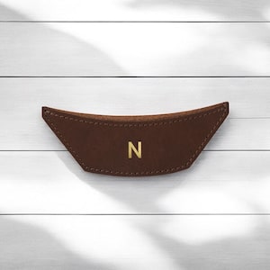 May include: A brown leather handle with a curved shape and a gold letter "N" in the center. The handle is stitched around the edges and appears to be made of high-quality leather. The handle is designed to be attached to a bag or other item.