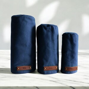 May include: Three navy blue cylindrical golf club head covers of varying heights, each featuring a rectangular brown leather patch at the base embossed with the letters BA and secured with two small metallic rivets on the left and right sides.