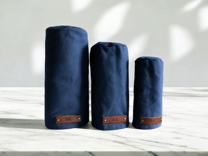 May include: Three navy blue cylindrical golf club head covers of varying heights, each featuring a rectangular brown leather patch at the base embossed with the letters BA and secured with two small metallic rivets on the left and right sides.