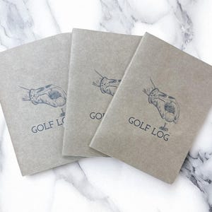 May include: Three gray notebooks with the words "GOLF LOG" printed on the cover. Each notebook features a black illustration of a gloved hand holding a golf ball. The notebooks are arranged on a white and gray marble surface.