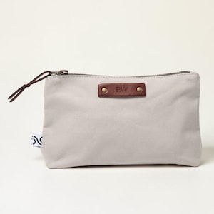 May include: A light gray canvas pouch with a brown leather accent and a zipper closure. The leather accent is embossed with the initials "PW" and secured with two rivets. A brown cord loop is attached to the zipper. A small tag with a black and white design is on the side.