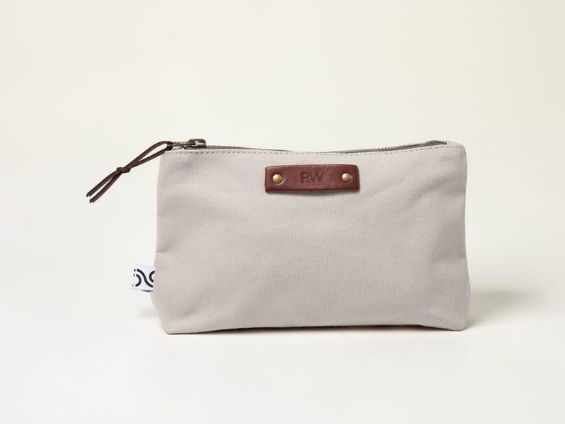 May include: A light gray canvas pouch with a brown leather accent and a zipper closure. The leather accent is embossed with the initials "PW" and secured with two rivets. A brown cord loop is attached to the zipper. A small tag with a black and white design is on the side.