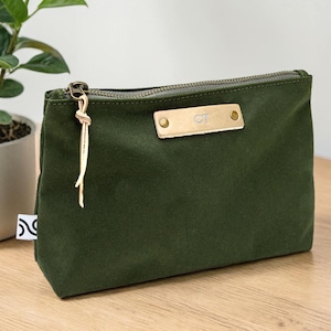May include: An olive green canvas zippered pouch with a gold-colored zipper and a leather tag. The tag is embossed with the initials "CT". A gold-colored tassel hangs from the zipper pull. The pouch is on a wooden surface.