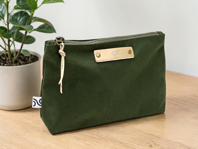 May include: An olive green canvas zippered pouch with a gold-colored zipper and a leather tag. The tag is embossed with the initials "CT". A gold-colored tassel hangs from the zipper pull. The pouch is on a wooden surface.