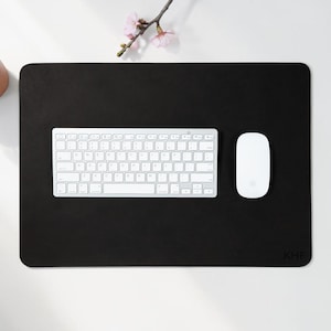 May include: A black rectangular desk mat with rounded corners, a white keyboard, and a white mouse. A small potted succulent and a sprig of pink flowers are in the upper left corner. The mat has the initials "KHF" in the lower right corner.