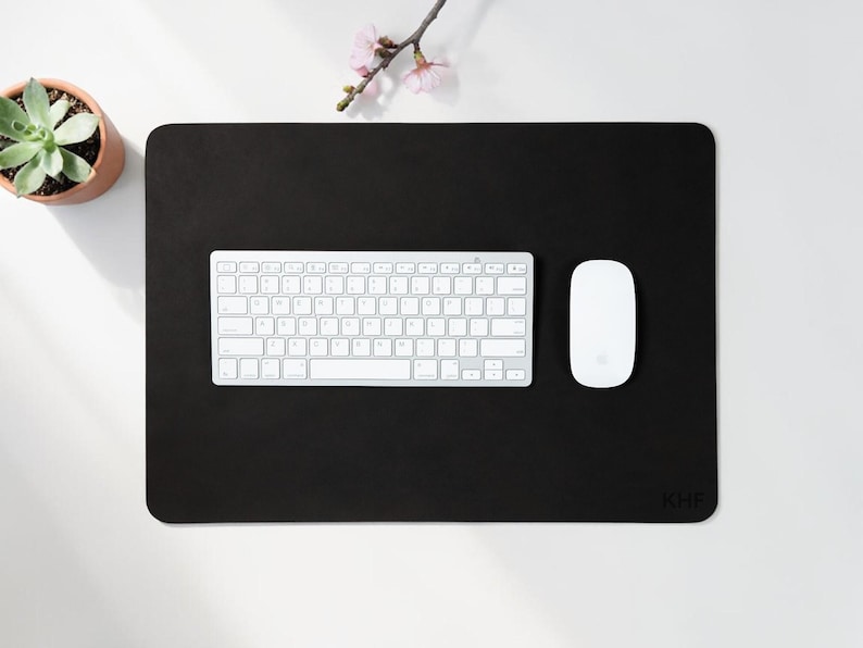 May include: A black rectangular desk mat with rounded corners, a white keyboard, and a white mouse. A small potted succulent and a sprig of pink flowers are in the upper left corner. The mat has the initials "KHF" in the lower right corner.