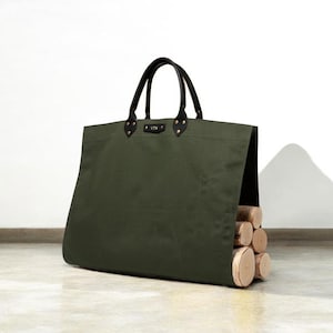May include: An olive green canvas log carrier with black leather handles and accents. The bag is filled with several cut logs, showcasing a practical and stylish design for firewood transport.