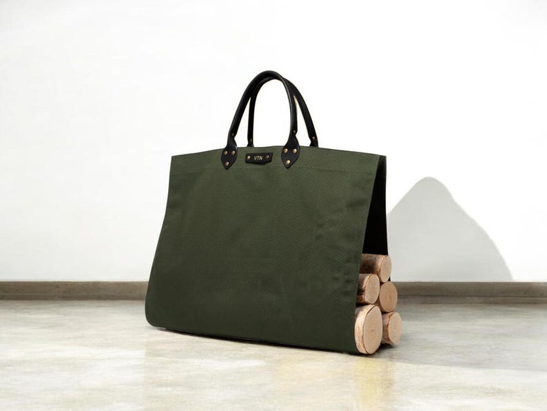 May include: An olive green canvas log carrier with black leather handles and accents. The bag is filled with several cut logs, showcasing a practical and stylish design for firewood transport.