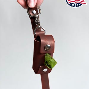 May include: A brown leather dog waste bag dispenser attached to a matching leather leash with a silver metal swivel snap hook and D-ring. The cylindrical holder features two silver snap buttons and contains a green plastic waste bag.