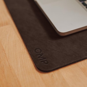 May include: A dark brown leather desk mat with the embossed letters "CMP" in the corner. A silver laptop rests on the mat, partially obscuring the surface. The mat is on a light wood surface.