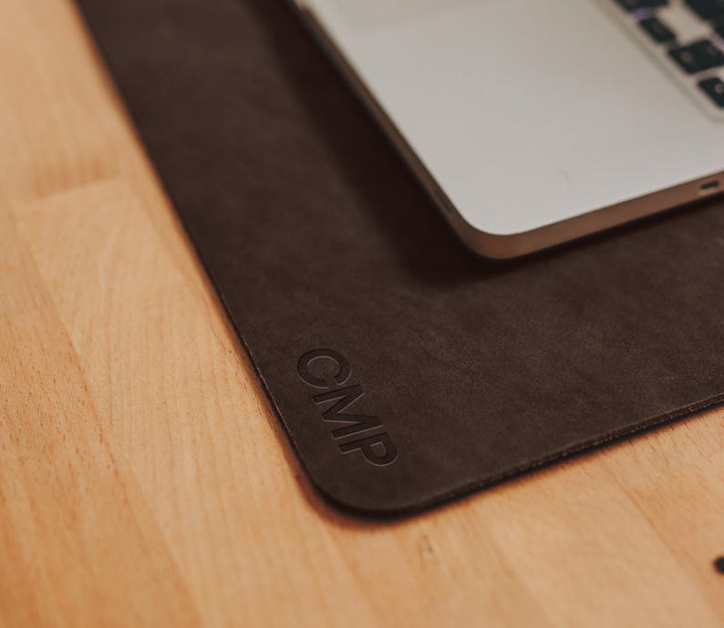 May include: A dark brown leather desk mat with the embossed letters "CMP" in the corner. A silver laptop rests on the mat, partially obscuring the surface. The mat is on a light wood surface.