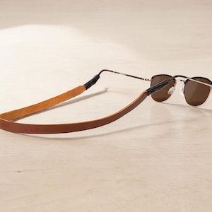 May include: Sunglasses with brown lenses and a thin, gold-colored frame, connected to a brown leather strap. The strap has the word "ANY" embossed on it. The sunglasses rest on a light-colored wooden surface.