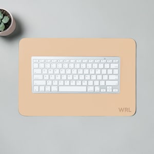 May include: A light beige rectangular desk mat with rounded corners, a silver keyboard, and the initials "WRL" in the bottom right corner. A small potted succulent is in the upper left corner. The keyboard is centered on the mat.