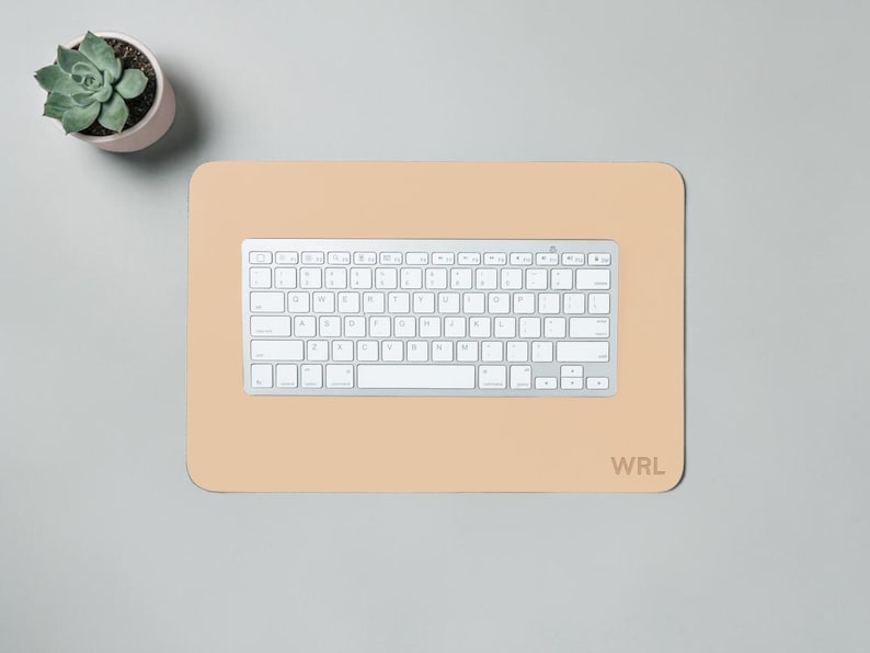 May include: A light beige rectangular desk mat with rounded corners, a silver keyboard, and the initials "WRL" in the bottom right corner. A small potted succulent is in the upper left corner. The keyboard is centered on the mat.