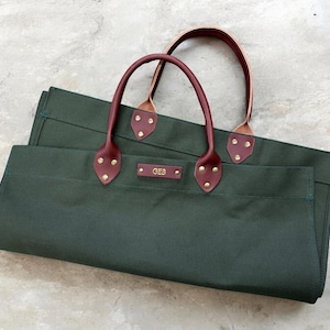 May include: Two dark green canvas tote bags with burgundy leather handles and accents. The handles are attached with brass rivets. The bags have a rectangular shape and a small rectangular tag with the initials "GEB".