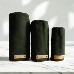 May include: Three dark olive green cylindrical golf club head covers of varying heights. Each cover features a rectangular tan leather patch at the base, secured with two brass rivets, and embossed with the letters DB in a simple sans-serif font.
