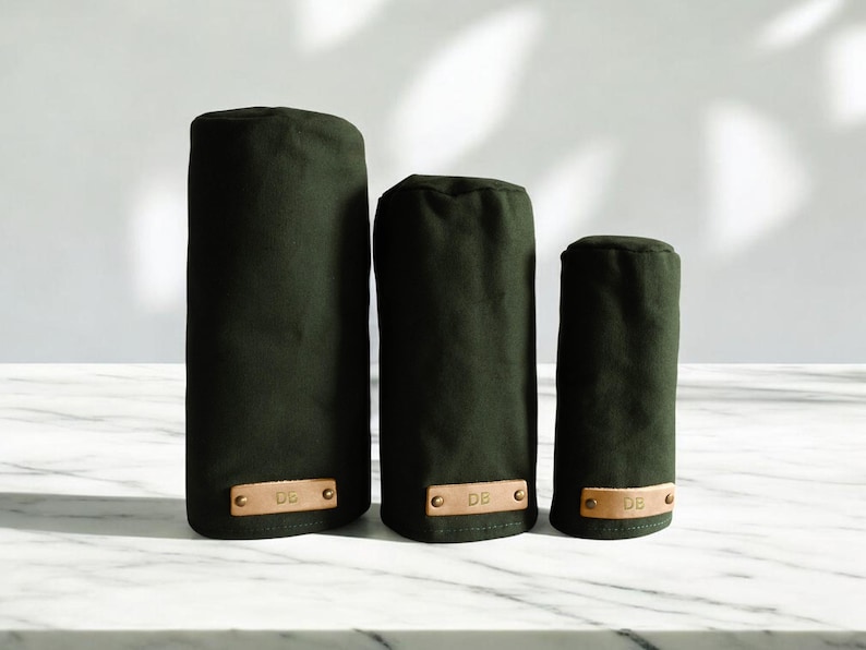 May include: Three dark olive green cylindrical golf club head covers of varying heights. Each cover features a rectangular tan leather patch at the base, secured with two brass rivets, and embossed with the letters DB in a simple sans-serif font.