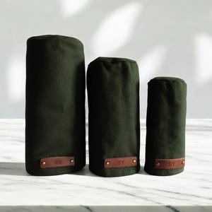 May include: Three dark olive green cylindrical golf club head covers of varying heights. Each cover features a rectangular brown leather patch at the base, embossed with the letters SY and secured with two small metallic rivets on each side.