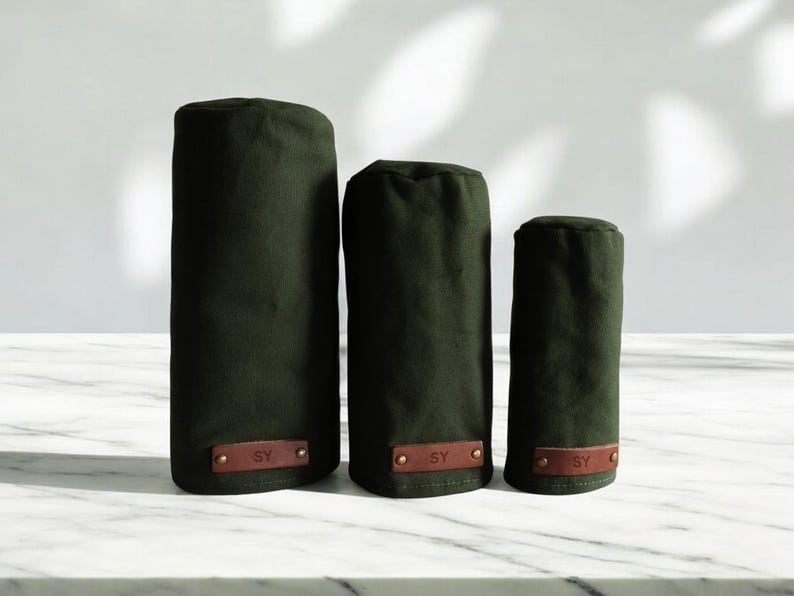 May include: Three dark olive green cylindrical golf club head covers of varying heights. Each cover features a rectangular brown leather patch at the base, embossed with the letters SY and secured with two small metallic rivets on each side.