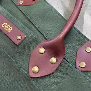May include: Detail of a green canvas tote bag with brown leather trim. The bag has a brown leather handle and a rectangular leather tag with the initials "GEB" in gold. Brass rivets are used to attach the leather.