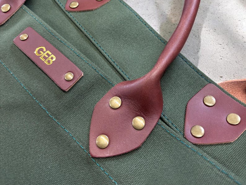 May include: Detail of a green canvas tote bag with brown leather trim. The bag has a brown leather handle and a rectangular leather tag with the initials "GEB" in gold. Brass rivets are used to attach the leather.