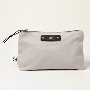 Könnte beinhalten: Eine hellgraue Canvas-Tasche mit schwarzem Reißverschluss und einem schwarzen Lederetikett. Das Etikett ist mit den Initialen "DF" geprägt und mit zwei goldenen Nieten befestigt. Eine schwarze Kordelschlaufe ist am Reißverschluss befestigt.