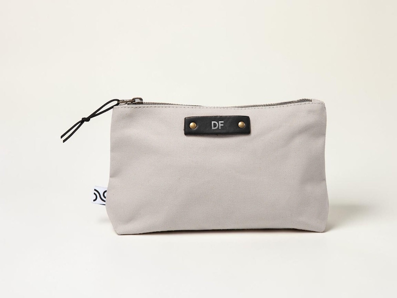May include: A light gray canvas pouch with a black zipper and a black leather tag. The tag is embossed with the initials "DF" and secured with two gold rivets. A black cord loop is attached to the zipper.