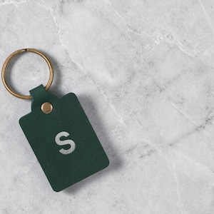 May include: A dark green leather keychain with a gold-colored ring and rivet. The keychain has a rectangular tag with the letter "S" in white. The keychain is on a white and gray marble surface.