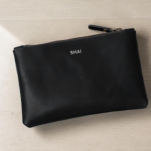 Pode incluir: Uma bolsa de couro preta com fecho de correr e o nome "SHAI" impresso em branco. A bolsa retangular est&aacute; sobre uma superf&iacute;cie de madeira clara. O puxador do z&iacute;per &eacute; preto.