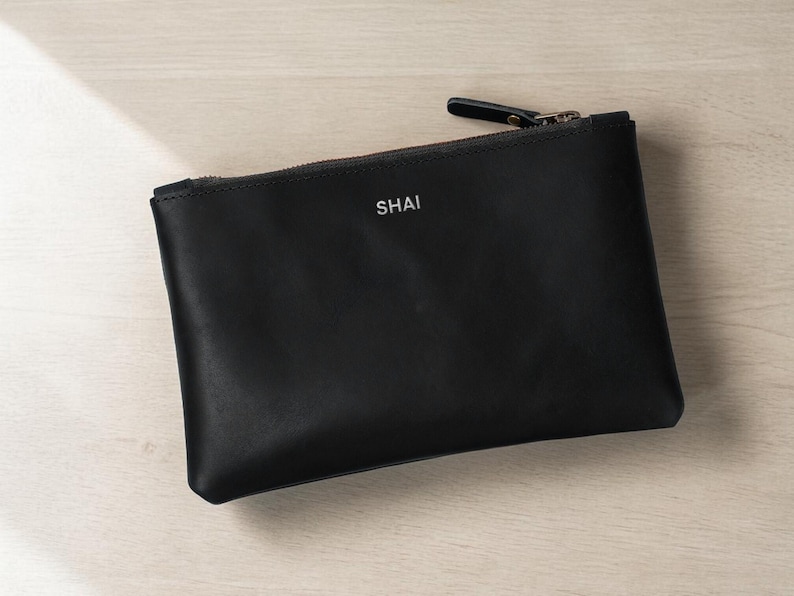 Pode incluir: Uma bolsa de couro preta com fecho de correr e o nome "SHAI" impresso em branco. A bolsa retangular est&aacute; sobre uma superf&iacute;cie de madeira clara. O puxador do z&iacute;per &eacute; preto.