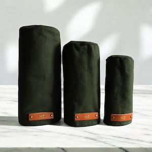 May include: Three dark olive green cylindrical golf club head covers of varying heights. Each cover features a rectangular tan leather patch at the base, secured with two brass rivets, and embossed with the letters NZ in a gold-colored font.